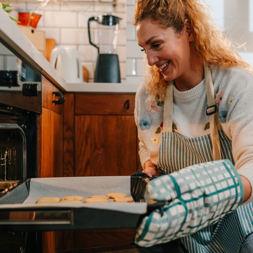 Happy woman wearing an apron and oven mitts, joyfully pulling a tray of freshly baked cookies from the oven in a sleek, modern kitchen filled with delightful aromas